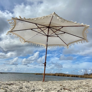 May include: A white crochet beach umbrella with a wooden pole and fringe trim, standing on a sandy beach with a body of water in the background.