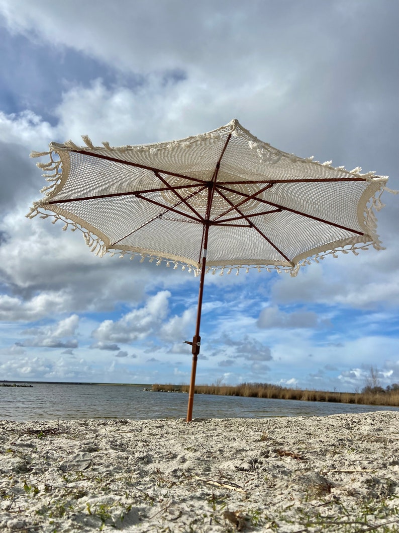 May include: A white crochet beach umbrella with a wooden pole and fringe trim, standing on a sandy beach with a body of water in the background.