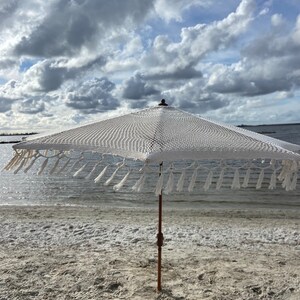 May include: A white crochet beach umbrella with fringe trim stands on a sandy beach with a body of water in the background. The sky is cloudy.
