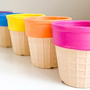 May include: Five colorful ice cream cone shaped planters in a row. The planters are painted in shades of purple, blue, yellow, orange, and pink. The planters are made of a textured material that resembles a waffle cone.