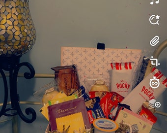 May include: A wicker basket filled with a variety of snacks and treats, including chocolate, cookies, and tea. The basket is decorated with a red and white striped ribbon.
