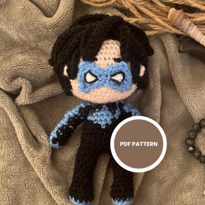 May include: A handmade Nightwing amigurumi doll with a blue mask and black suit. The doll is crocheted and has a round white sticker that says "PDF PATTERN". The doll is on a gray blanket with other decorative items.