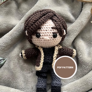 Könnte beinhalten: Handgefertigte Häkelpuppe mit braunen Haaren, einer braunen Jacke mit beigen Verzierungen und grauen Hosen. Die Puppe hat einen neutralen Hautton und liegt auf einer grauen, weichen Oberfläche. Ein runder Aufkleber mit dem Text "PDF PATTERN" ist sichtbar.