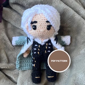 以下が含まれることがあります： 白い髪の毛で、黒と金の衣装と薄緑と白のコートを着た手作りのかぎ針編み人形。人形は黒い目の丸い顔をしており、灰色の毛布の上にあります。「PDF PATTERN」と書かれた円形のグラフィック。