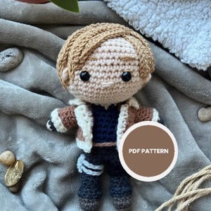 Puede incluir: Muñeca amigurumi de crochet con cabello castaño, una chaqueta marrón y blanca y pantalones negros. La muñeca sostiene un círculo marrón con el texto "PDF PATTERN" en blanco.
