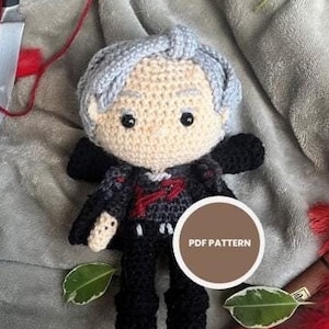 May include: Crocheted amigurumi doll with gray hair, black clothing, and a red heart on its chest. The doll is sitting on a gray blanket with red feathers and green leaves. The text "PDF PATTERN" is on a brown circle.