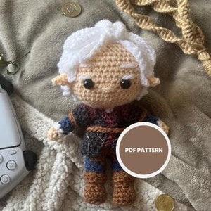 May include: Crocheted amigurumi doll with white hair, brown eyes, and a burgundy and black outfit. The doll is sitting on a white fluffy blanket with a brown tag that says "PDF PATTERN".