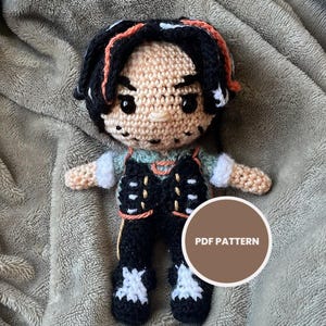 以下が含まれることがあります： 黒髪とオレンジ色のハイライトが入った手作りのかぎ針編み人形。黒いベストと白いシャツを着ています。人形は灰色の毛布の上にあり、「PDF PATTERN」のラベルが付いています。