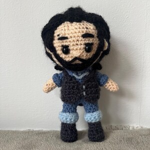 May include: Crocheted amigurumi doll with black hair, a black vest, blue jeans, and black boots. The doll has a brown face with black eyes and a black beard.