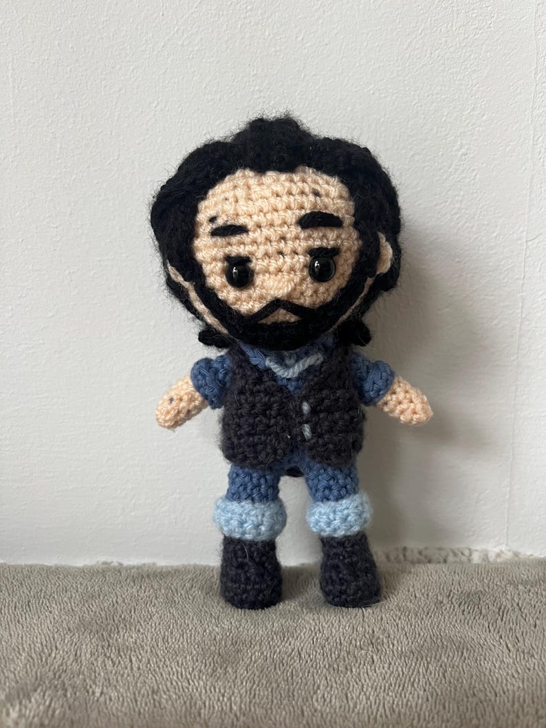 May include: Crocheted amigurumi doll with black hair, a black vest, blue jeans, and black boots. The doll has a brown face with black eyes and a black beard.