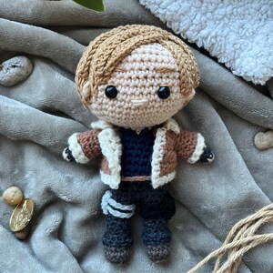 May include: Crocheted doll with brown hair, a brown jacket with a white fur collar, a blue shirt, and black pants. The doll is wearing a white and gray bandage on its left leg.