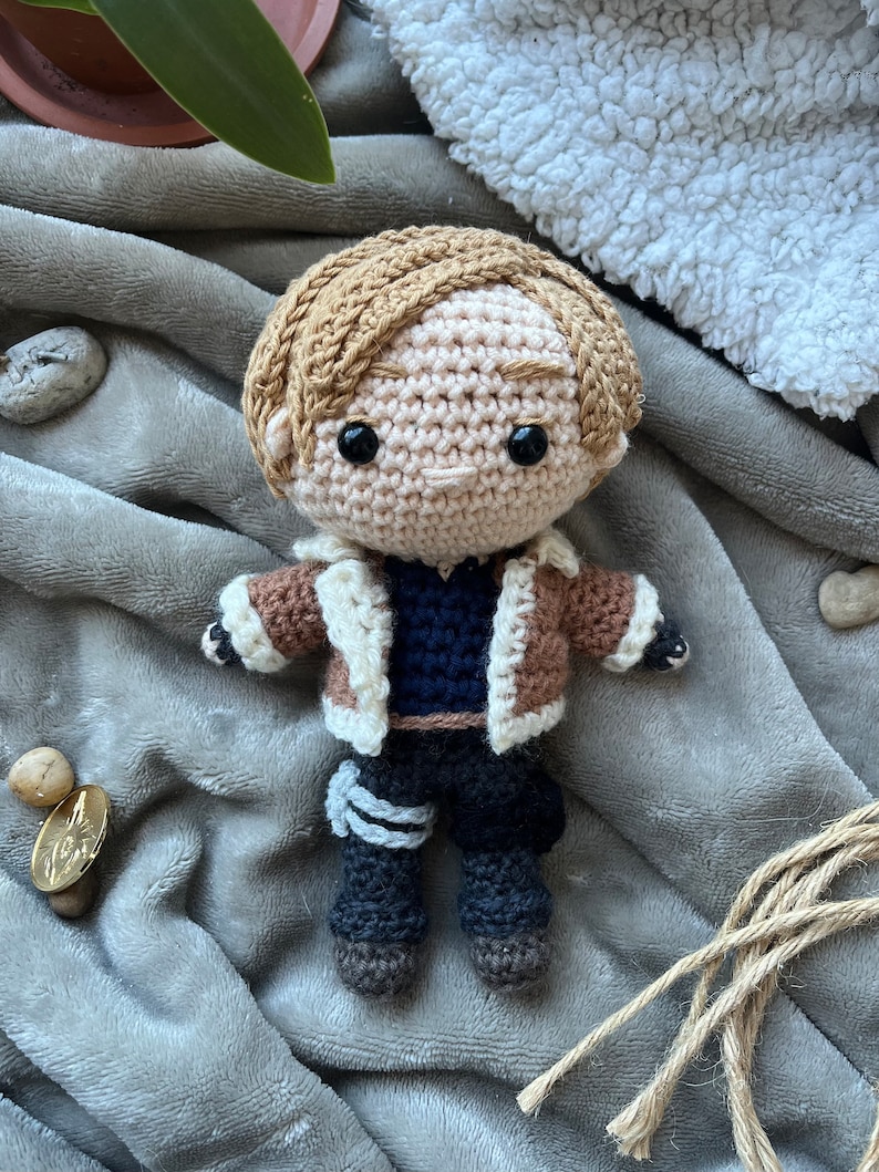 May include: Crocheted doll with brown hair, a brown jacket with a white fur collar, a blue shirt, and black pants. The doll is wearing a white and gray bandage on its left leg.