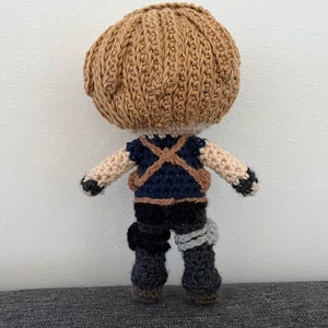 May include: A crocheted doll with brown hair, a blue and brown vest, and black pants. The doll is wearing a grey bandage on its right leg.