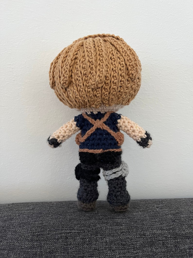 May include: A crocheted doll with brown hair, a blue and brown vest, and black pants. The doll is wearing a grey bandage on its right leg.