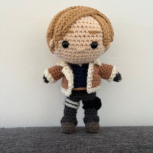 May include: A crocheted doll with brown hair, a brown jacket, and black pants. The doll has a neutral expression and black eyes.