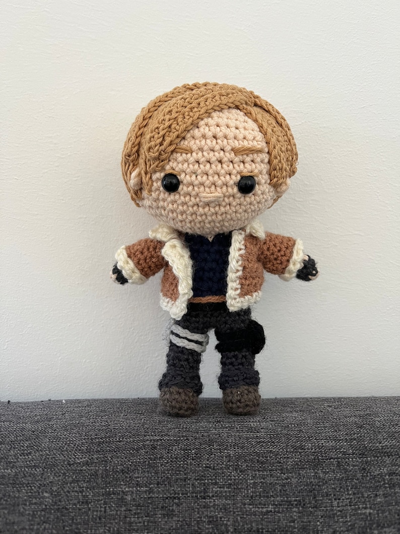 May include: A crocheted doll with brown hair, a brown jacket, and black pants. The doll has a neutral expression and black eyes.