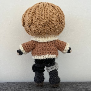 May include: A crocheted doll with brown hair, a brown jacket with white trim, black pants, and gray leg warmers.