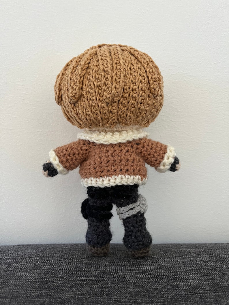 May include: A crocheted doll with brown hair, a brown jacket with white trim, black pants, and gray leg warmers.