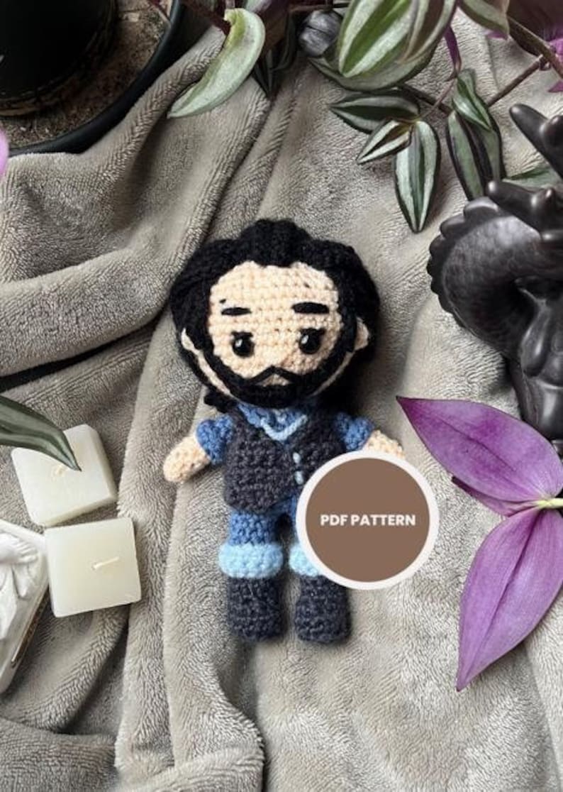 May include: Crocheted amigurumi doll of a person with black hair, a black vest, blue pants, and brown boots. The doll is sitting on a gray blanket with a purple leaf and a brown circle with the text "PDF PATTERN" on it.
