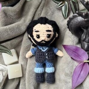 May include: A crocheted plush toy of a person with black hair, a black vest, blue jeans, and black shoes. The toy is sitting on a gray blanket.