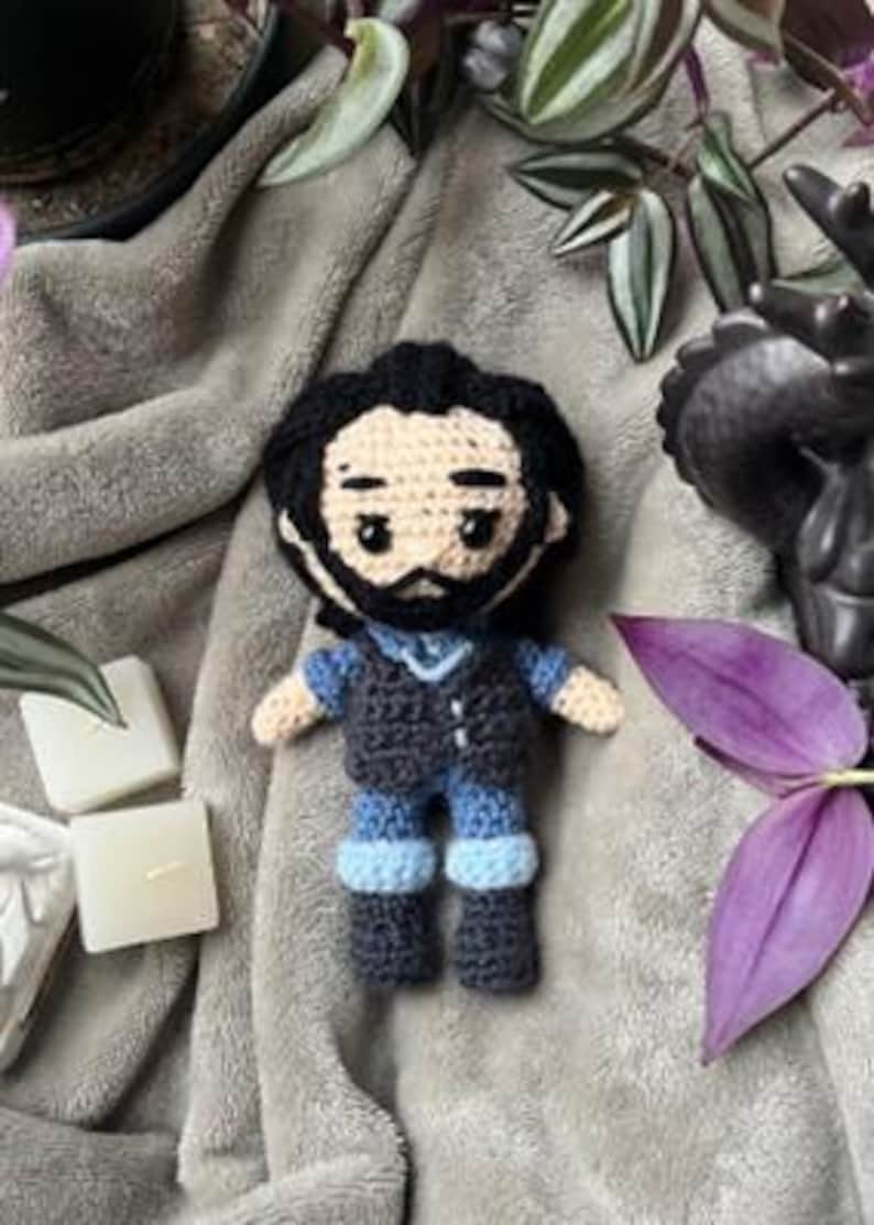 May include: A crocheted plush toy of a person with black hair, a black vest, blue jeans, and black shoes. The toy is sitting on a gray blanket.