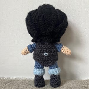 May include: A crocheted doll with black hair, a dark blue shirt, and blue and white pants. The doll is wearing black boots.