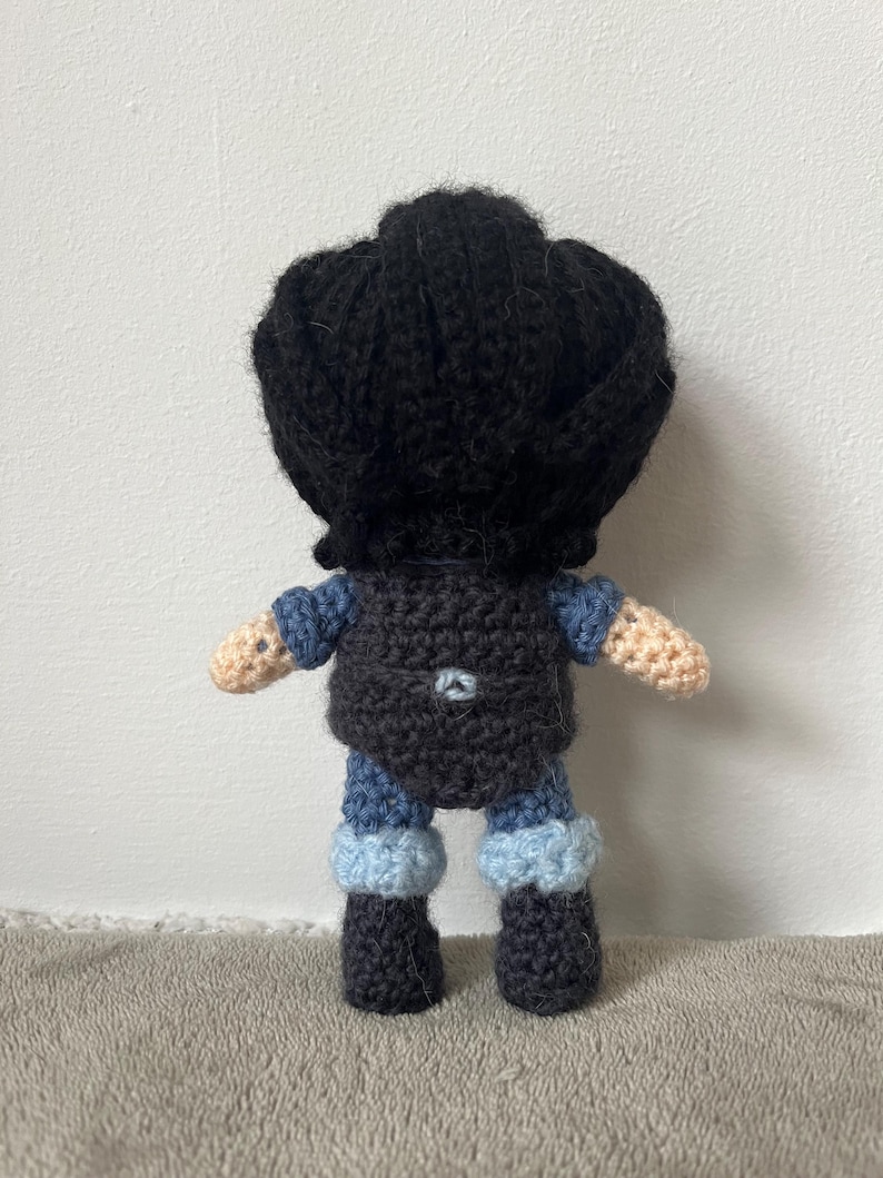 May include: A crocheted doll with black hair, a dark blue shirt, and blue and white pants. The doll is wearing black boots.