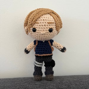 May include: A crocheted doll with brown hair, a blue shirt, and gray pants. The doll is wearing a backpack and has a friendly expression.