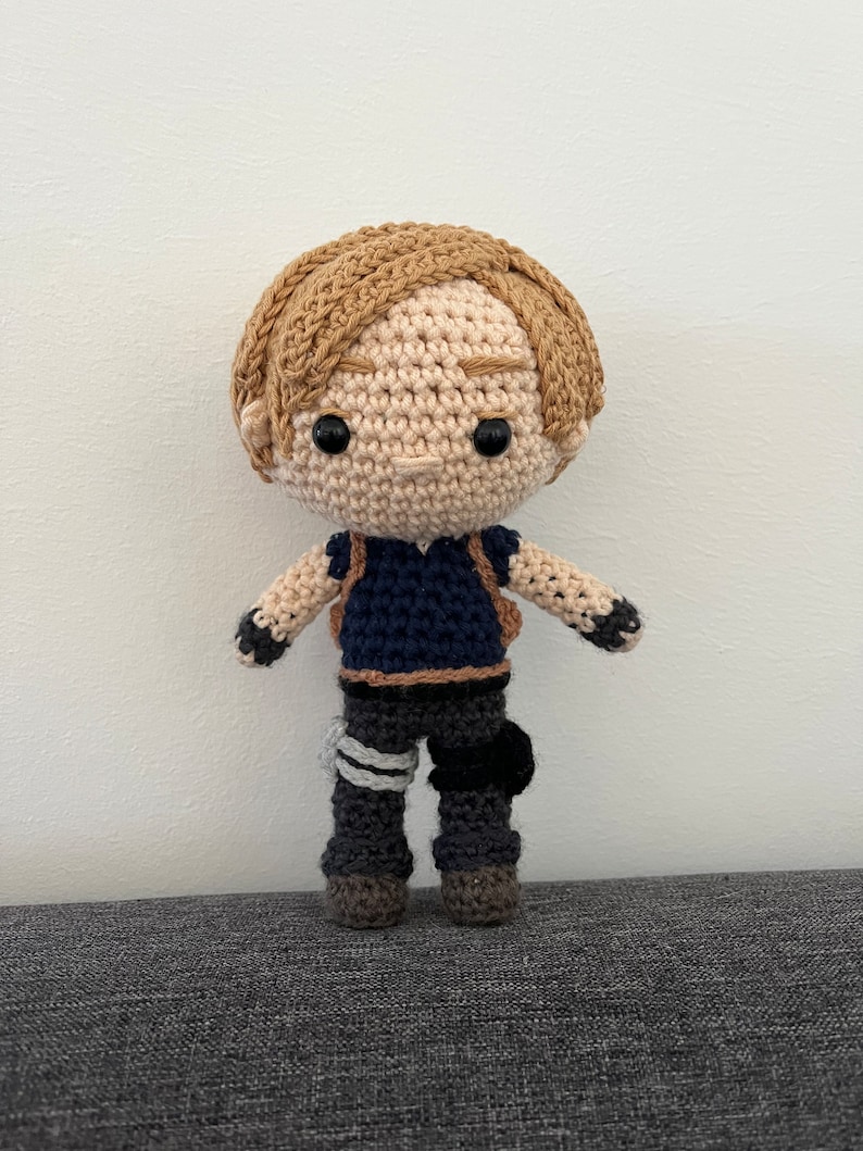 May include: A crocheted doll with brown hair, a blue shirt, and gray pants. The doll is wearing a backpack and has a friendly expression.