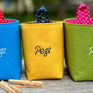 May include: Three colorful fabric containers for pegs, each with a contrasting polka dot lining and a handle. The containers are blue, yellow, and green, with the word "Pegs" embroidered in white. Wooden clothespins are visible.