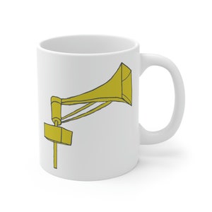 May include: White ceramic mug featuring a yellow illustration of a vintage megaphone. The megaphone design is outlined in black and includes a rectangular base and a horn shape. The mug has a curved handle and a glossy finish.