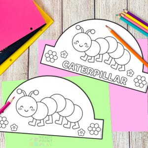 May include: Two black and white caterpillar-themed paper crowns with the word "CATERPILLAR" printed on them. The crowns are on pink and green paper, with colored pencils and folders in the background. The crowns have a smiling caterpillar design.