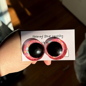 May include: Two pink googly eyes with black pupils and iridescent glitter. The eyes are on a white card with the text "Hooray! Your squishy item is"