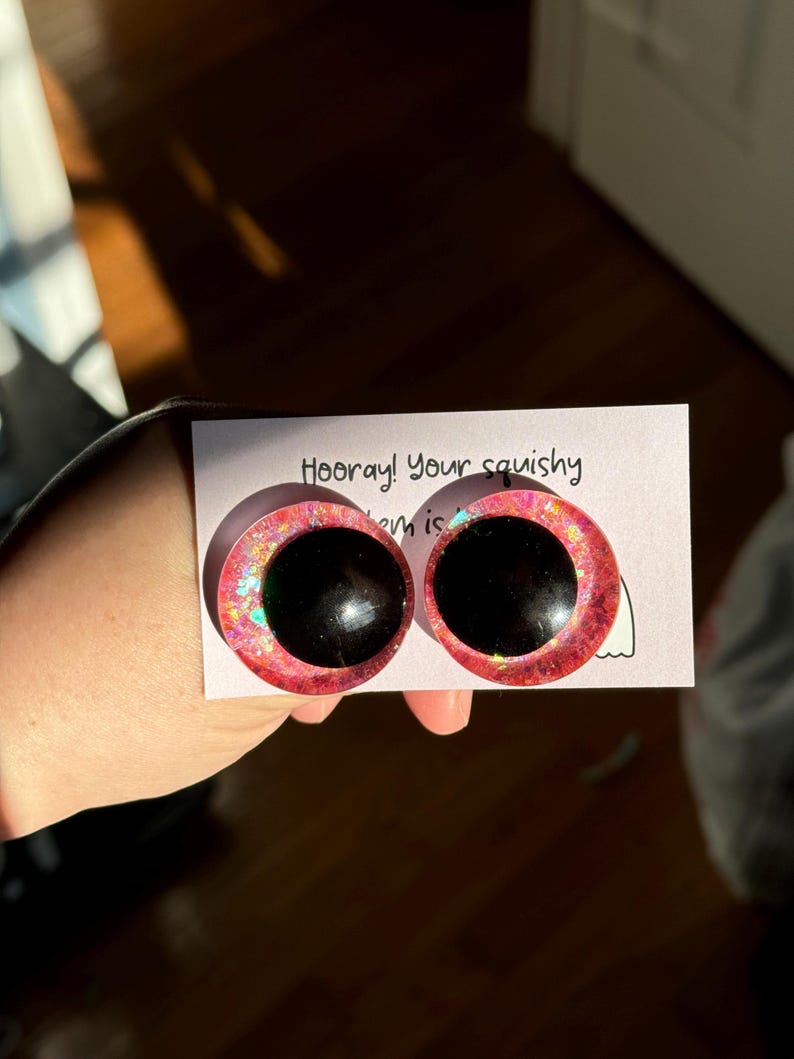 May include: Two pink googly eyes with black pupils and iridescent glitter. The eyes are on a white card with the text "Hooray! Your squishy item is"