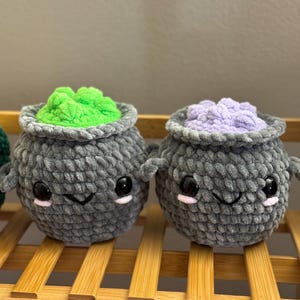 May include: Two gray crocheted cauldron plushies with cute faces. One cauldron has green yarn inside and the other has purple yarn inside.