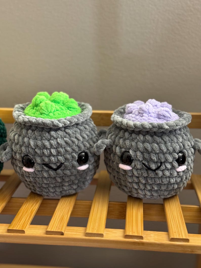 May include: Two gray crocheted cauldron plushies with cute faces. One cauldron has green yarn inside and the other has purple yarn inside.