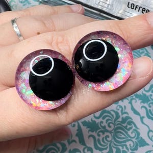 May include: Two large pink and iridescent glitter googly eyes with black pupils and white outlines.