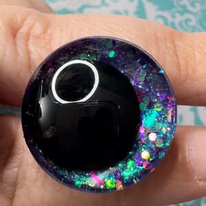 May include: A black and iridescent glitter resin eye with a white circle in the center. The eye is approximately 1 inch in diameter.