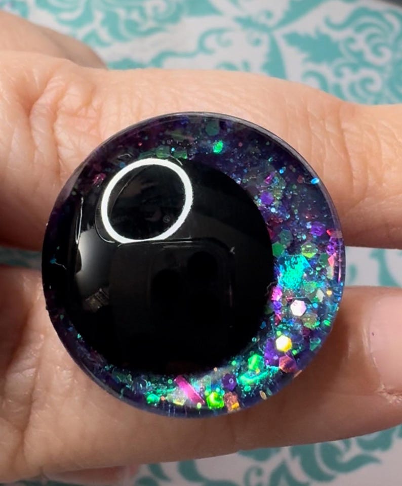 May include: A black and iridescent glitter resin eye with a white circle in the center. The eye is approximately 1 inch in diameter.