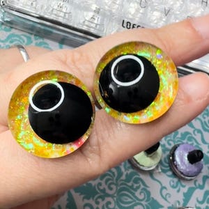 May include: Two yellow and gold glitter safety eyes with black pupils and white outlines. The eyes are on a hand, and there are other safety eyes in the background.