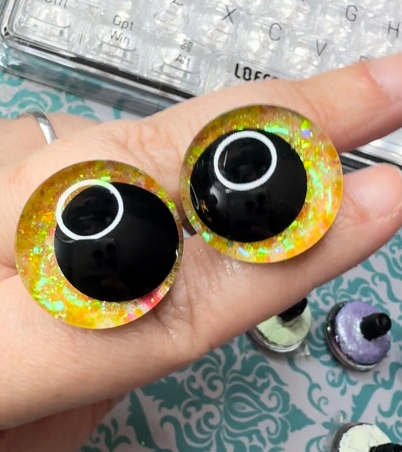 May include: Two yellow and gold glitter safety eyes with black pupils and white outlines. The eyes are on a hand, and there are other safety eyes in the background.