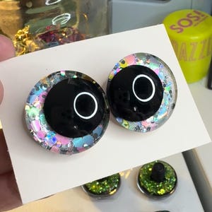 May include: Two black plastic eyes with a white ring around the pupil. The eyes are surrounded by iridescent glitter.