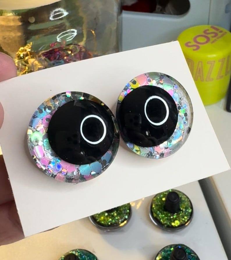 May include: Two black plastic eyes with a white ring around the pupil. The eyes are surrounded by iridescent glitter.