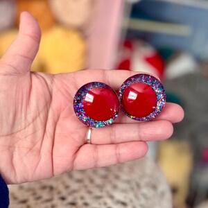 May include: Two red round resin buttons with a glittery purple, blue, and green border.