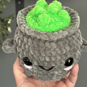 May include: A gray crocheted cauldron with a green gooey substance inside. The cauldron has a cute cartoon face with black eyes and a small smile.