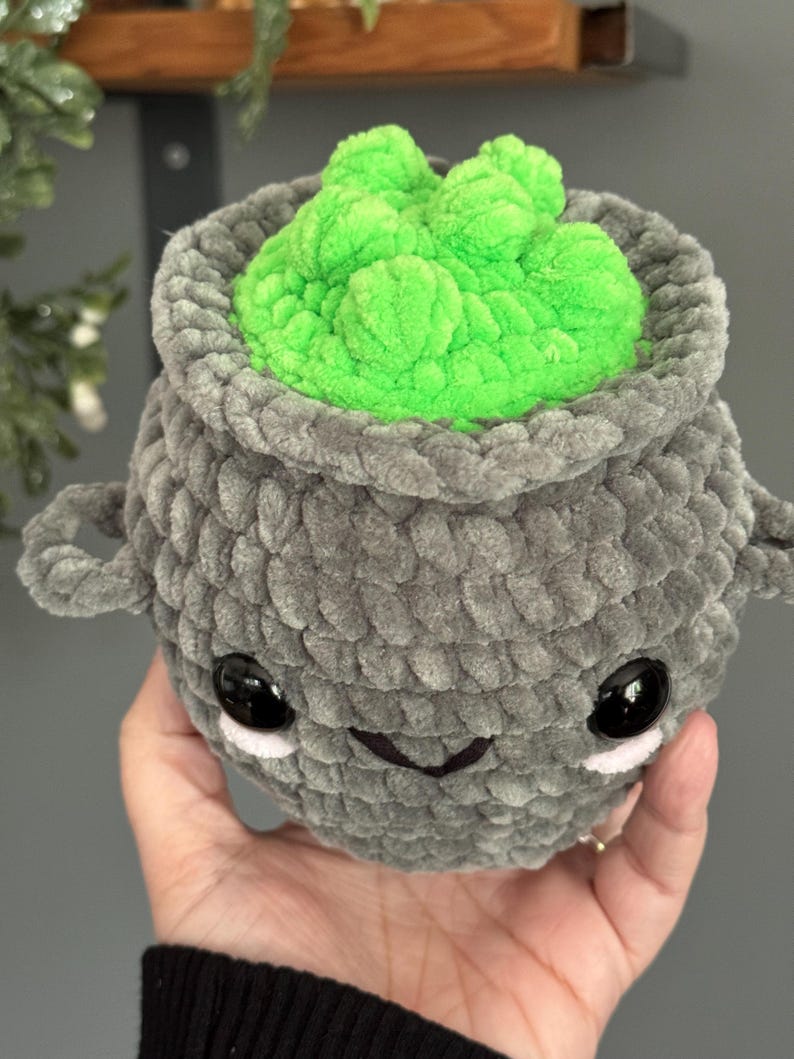 May include: A gray crocheted cauldron with a green gooey substance inside. The cauldron has a cute cartoon face with black eyes and a small smile.