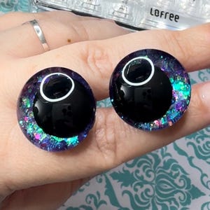 May include: Two black and white plastic eyes with a purple, blue, and green glitter rim. The eyes are on a hand.