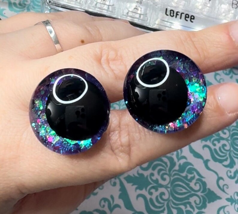 May include: Two black and white plastic eyes with a purple, blue, and green glitter rim. The eyes are on a hand.