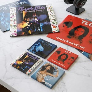 May include: A collection of square coasters featuring album art from artists like Prince, TLC, Mary J. Blige, and Beyoncé. The coasters have vibrant colors and images, including the album "Purple Rain" and "CrazySexyCool". Black headphones rest on a stand.