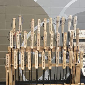 May include: A wooden display rack holding multiple beer tap handles. The handles are made of wood and metal, with various logos and text, including "Happy Camper," "Fire King," and "Packers." The rack is on wheels.